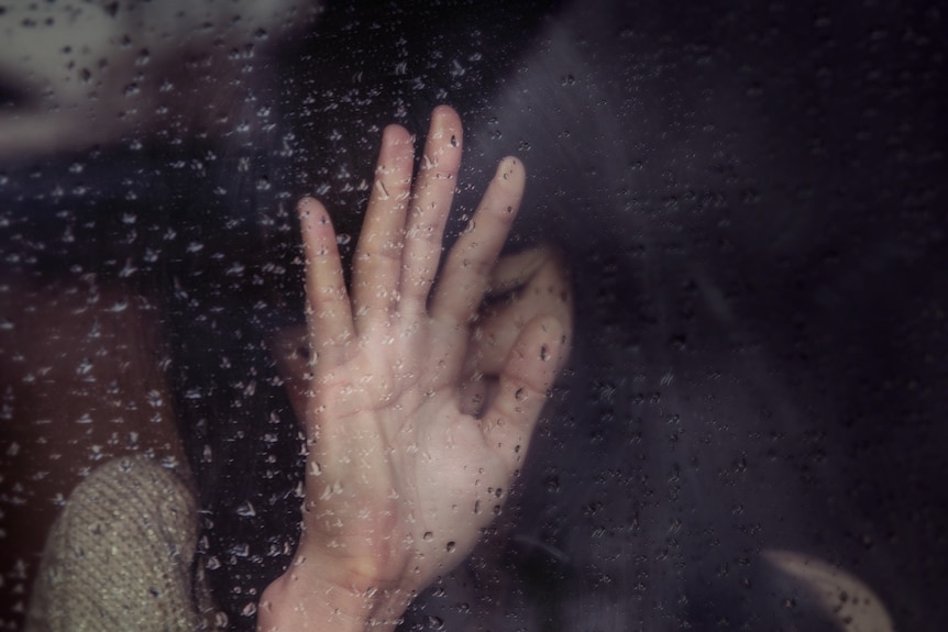 A Distressed woman with her hand on a pane of glass with rain droplets.