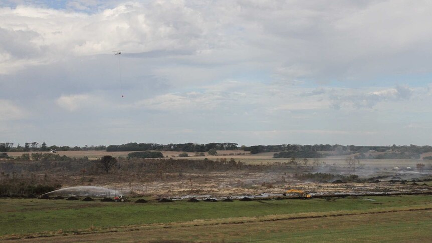 Water is sprayed on the peat fire, while an excavator digs a trench around the area. A helicopter flies overhead.