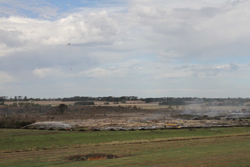 Water is sprayed on the peat fire, while an excavator digs a trench around the area. A helicopter flies overhead.