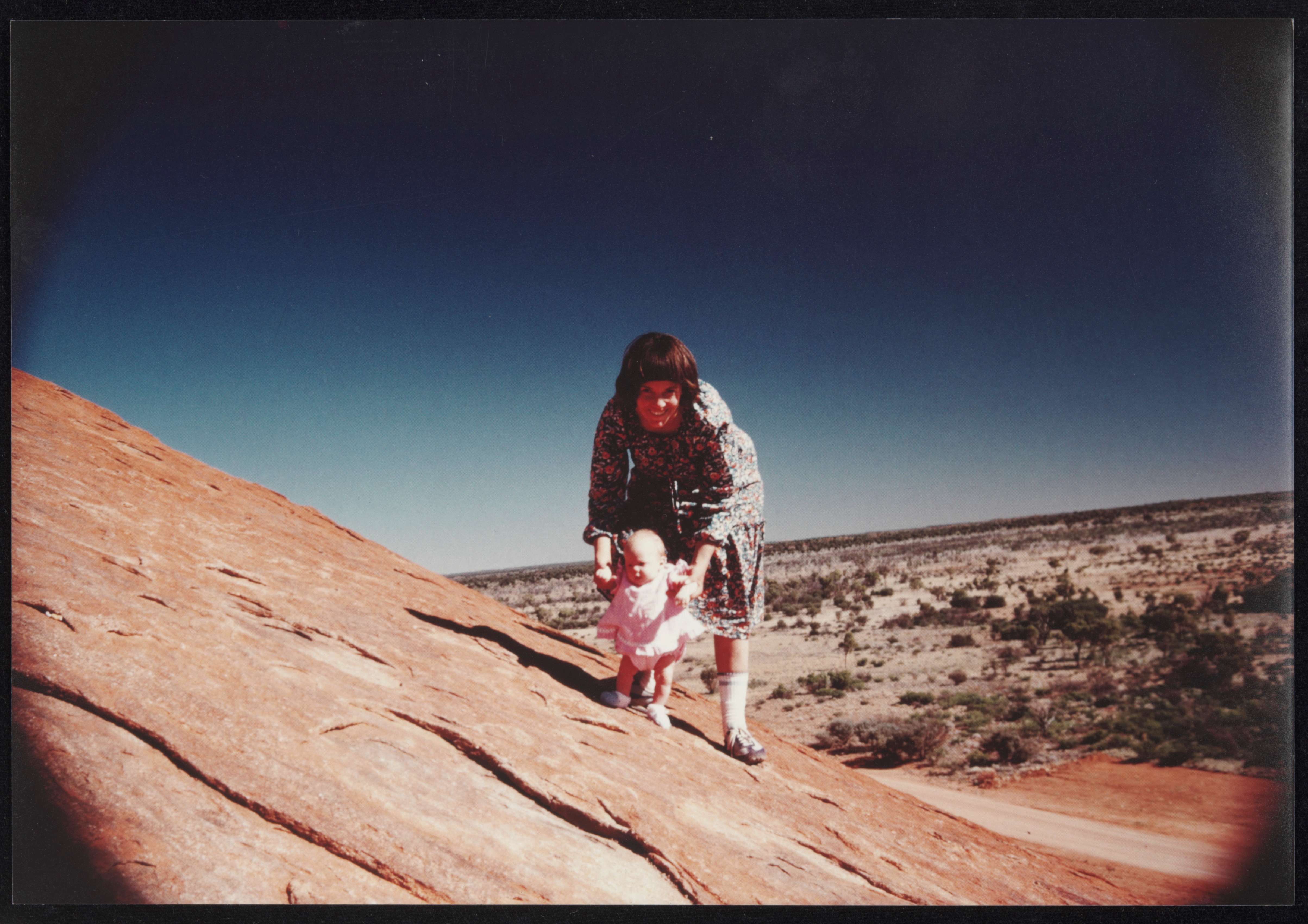 An older photo shows a mother with her infant daughter standing on a red rock in the outback, the baby girl is wearing pink.