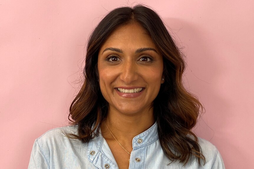 A woman in a blue shirt smiles into the camera, she's standing in front of a pink wall 