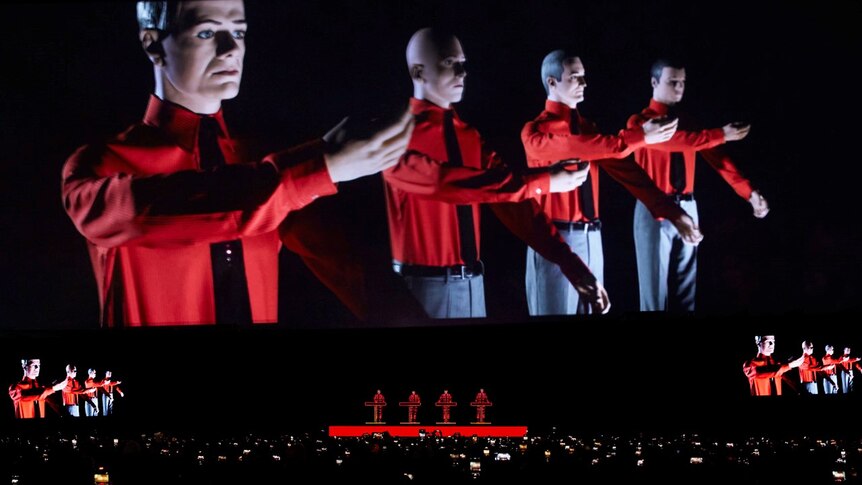 four members of Kraftwerk perform live, dwarfed by a giant projection of four robotic members in red shirts and grey pants