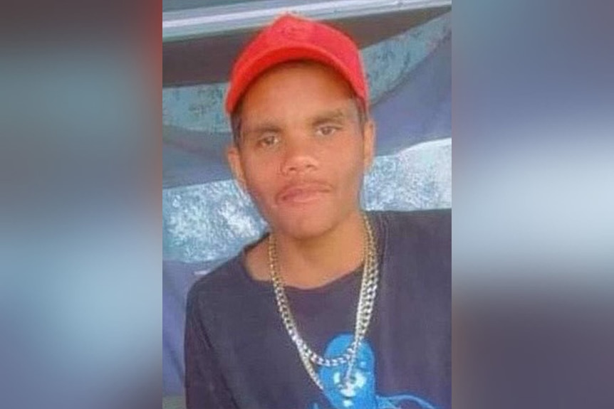 A close-up head and shoulders shot of a teenage Indigenous boy wearing a red cap, blue shirt and gold chains.