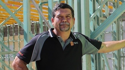 a man wearing a black polo and greying goatee leans against a steel frame