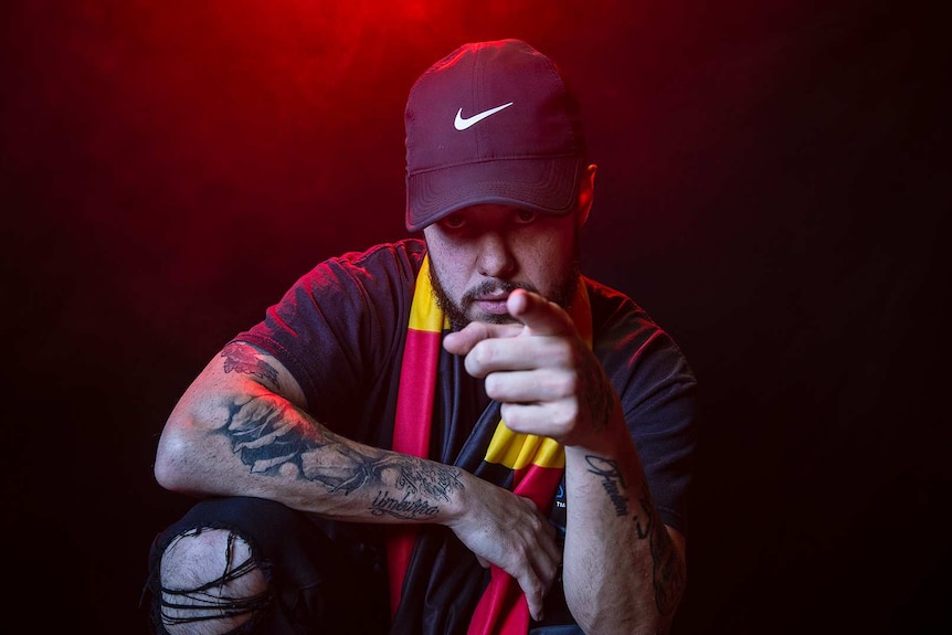 Colour portrait of rapper Nooky in front of black background with red smoke and pointing to camera.