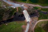 A bird's eye view of a concrete crossing over a small creek on green farmland lined with trees.