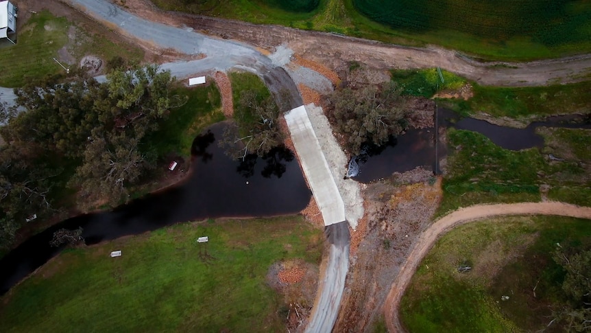 A bird's eye view of a concrete crossing over a small creek on green farmland lined with trees.