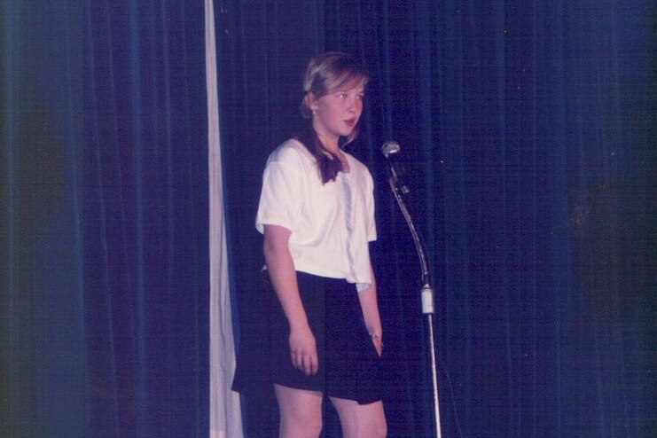 A girl stands on the stage at a microphone speaking, with the curtains drawn behind her