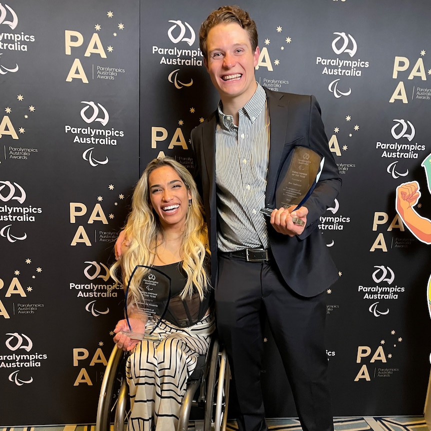 A woman in a wheelchair smiles and holds an award, a man stands next to her, holding an award, smiling.