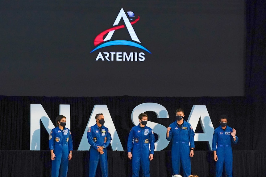 A group of five people stand on a stage in blue NASA uniforms