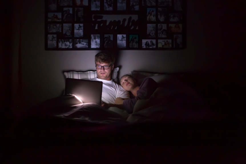 Middle-aged blond man with glasses is working on his laptop in bed at night while his female partner is asleep next to him. 
