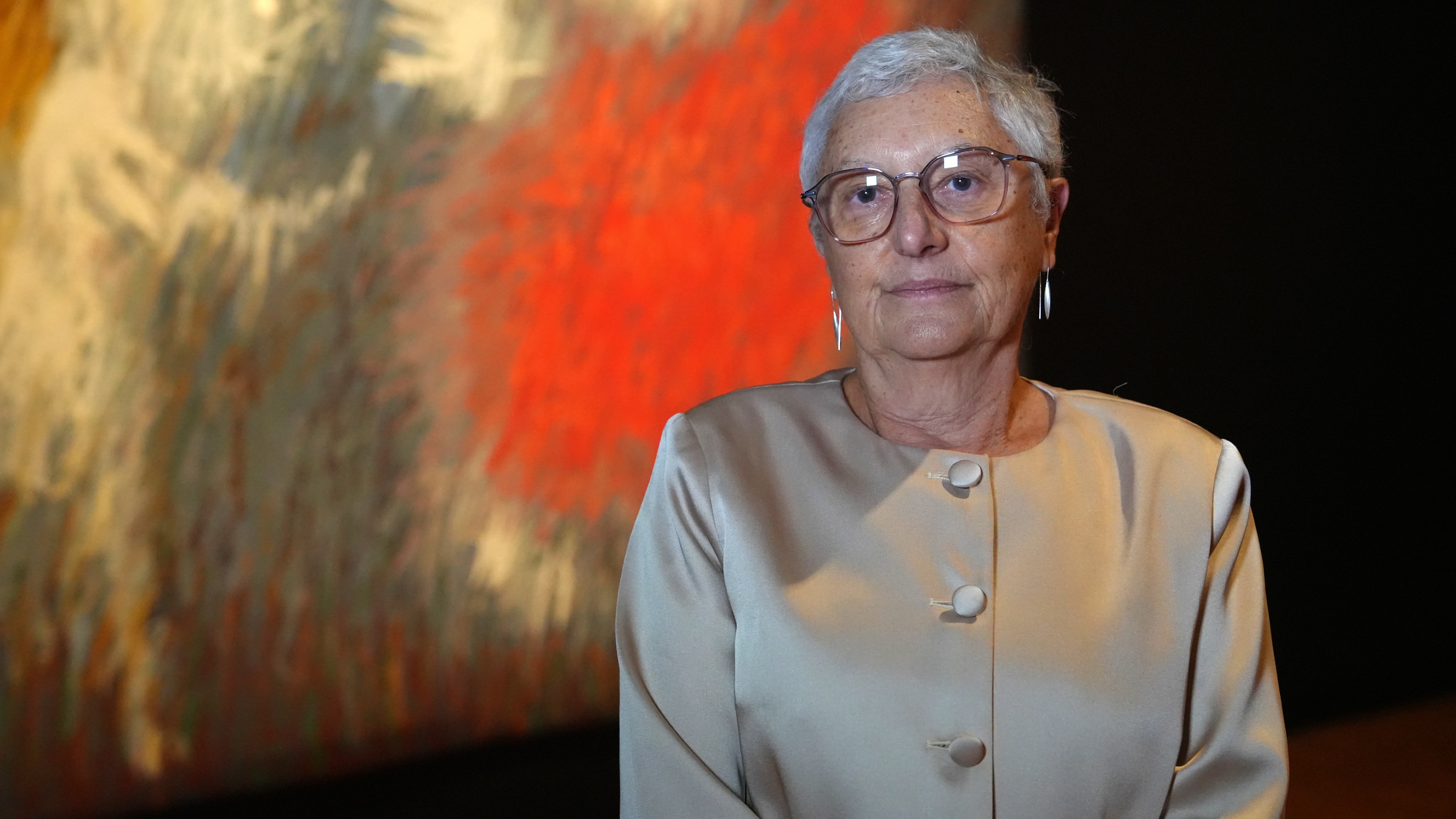 A woman with short grey hair and glasses stands in an art gallery.