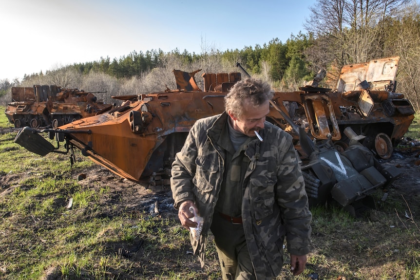 A man in a military style jacket stands near two destroyed armoured vehicles.