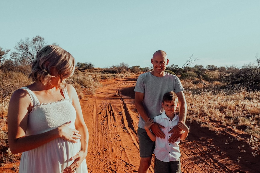 Louise Sylvia holding pregnant stomach with partner and child in back in the outback