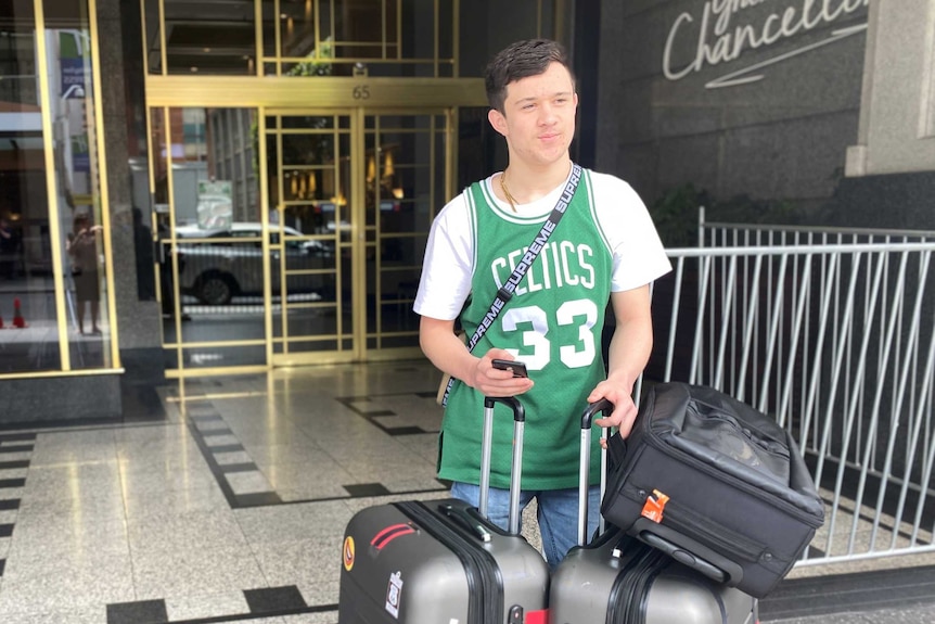 A man wearing a basketball shirt stands outside a hotel lobby with three suitcases