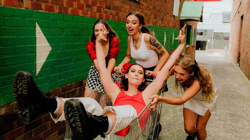 four young women from the band "Teen Jesus and the Jean Teasers" ride in a shopping trolley through a city alleyway