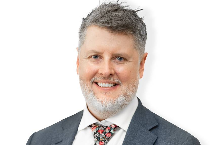 A man with grey hair and beard wearing a suit and loose floral tie looks at the camera