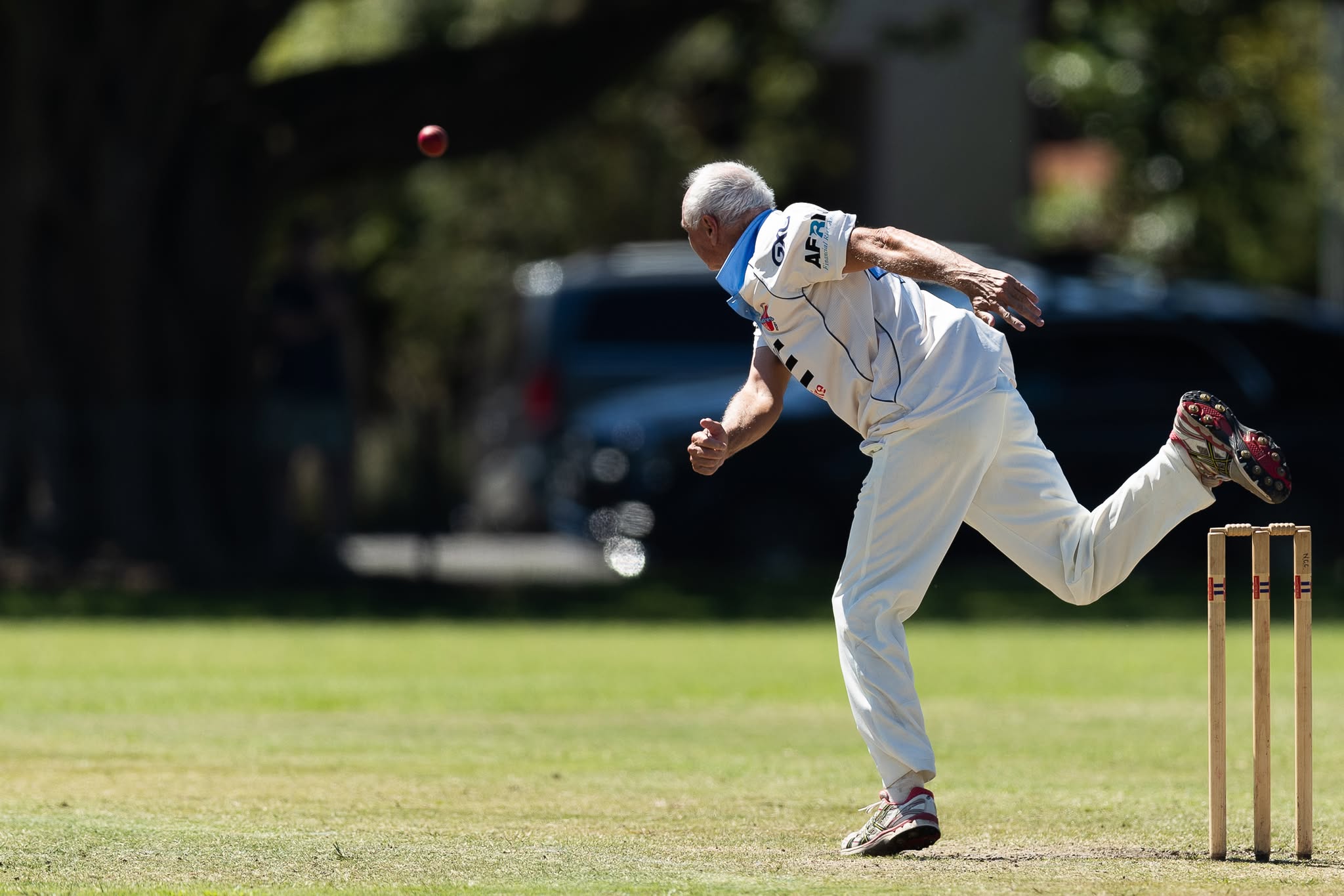 A man in cricket whites, with white hair, bowls a ball on a cricket field.