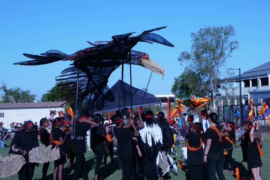 A group of children wearing black clothes and red headbands hold up a giant puppet of a bird.