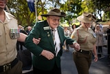an old man wearing a hat being held under his arms by a man and a woman wearing green uniforms