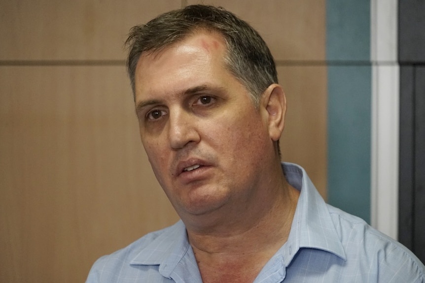 A man in a blue collared shirt stands in an office and looks concerned.