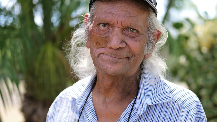 An elderly Indigenous man wearing a patterned button-up shirt and cap, smiling with some trees behind him.