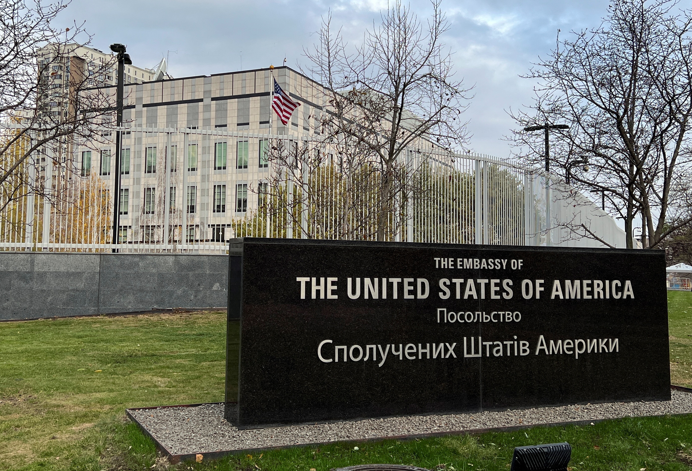 A black stone sign reading 'The United States of America' in English and Ukrainian in front of a silver fence and pale building