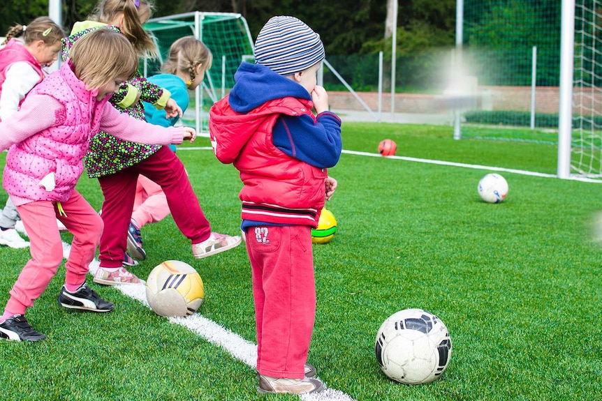 Small children kick soccer balls toward a goal, while one child stands off to the side, too tentative to have a kick.