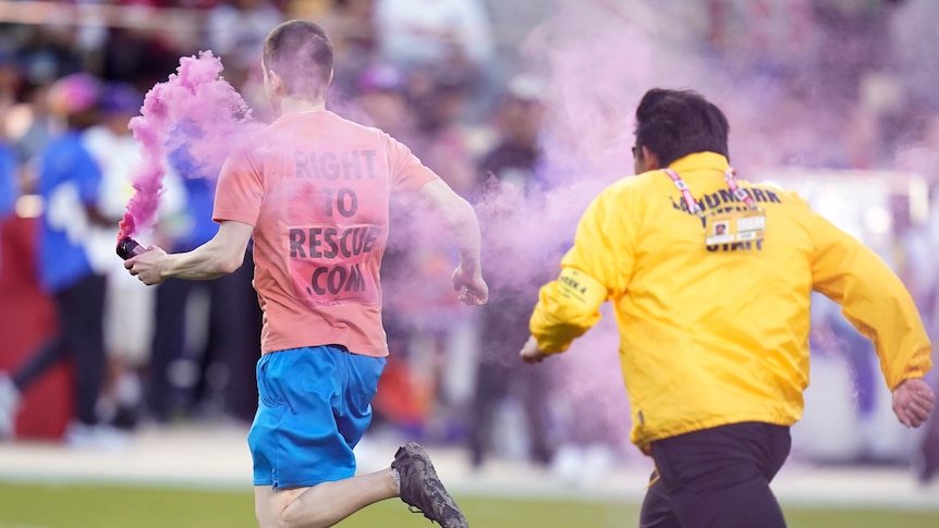 A man wearing a T-shirt and shorts and carrying a pink smoke flare runs across a field being chased by security during NFL game.