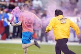 A man wearing a T-shirt and shorts and carrying a pink smoke flare runs across a field being chased by security during NFL game.