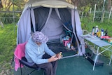 Man sitting in a folding chair in front of a tent with a cap on his head obscuring his face.