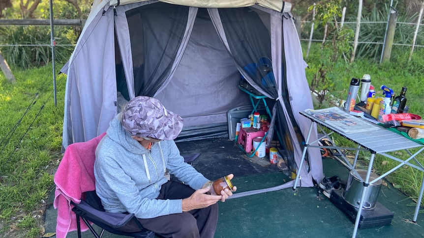 Man sitting in a folding chair in front of a tent with a cap on his head obscuring his face.