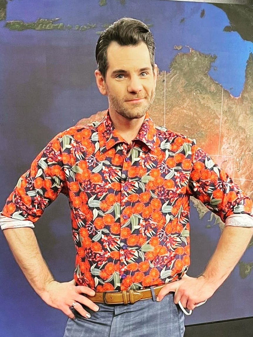 A man in a colourful patterned red shirt stands with hands on hips in front of weather map on television set.