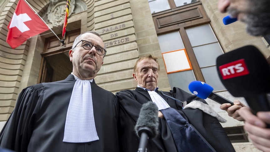 Two older white man in legal garb stand outside a courthouse in front of a bank of microphones.