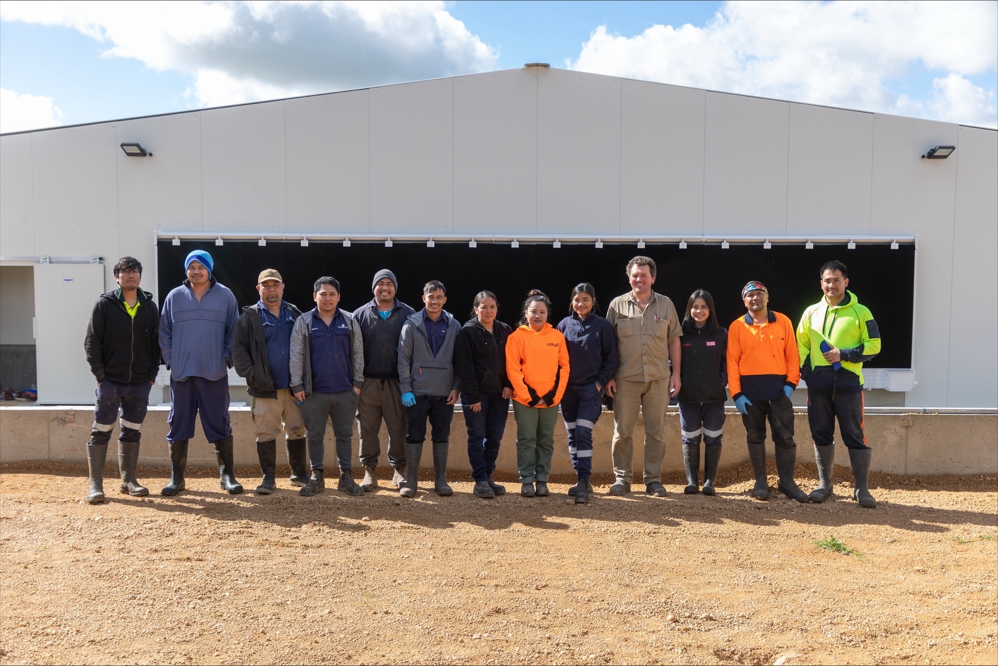 team of thirteen stand outside shed smiling
