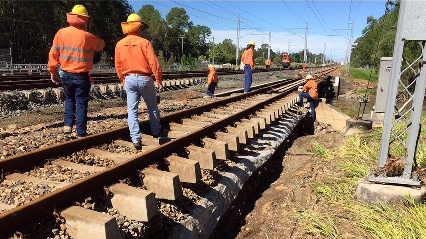 Workers in orange shirts, denim jeans and hard hats inspect rail lines.