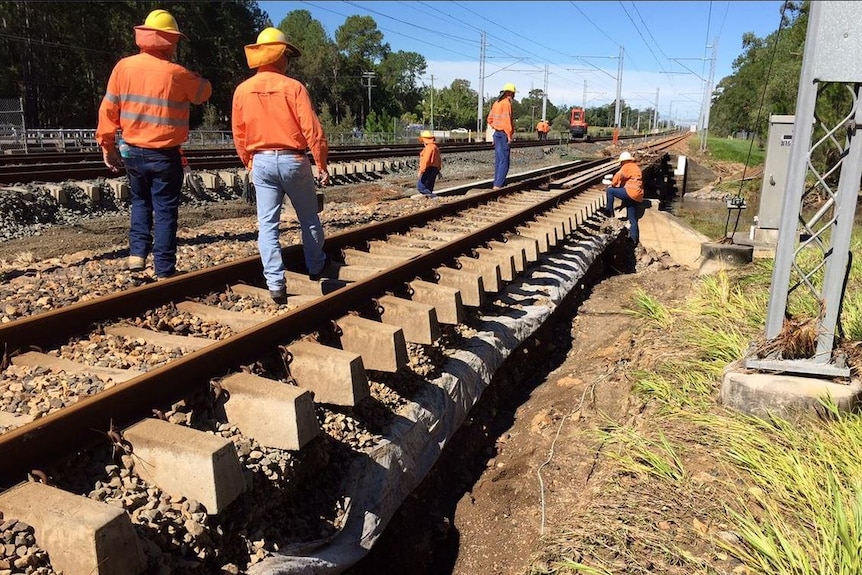 Workers in orange shirts, denim jeans and hard hats inspect rail lines.