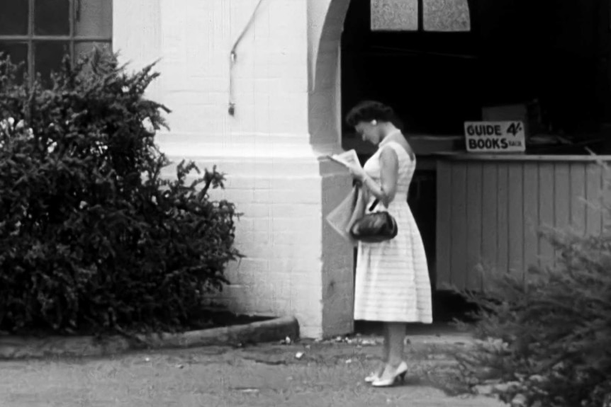 A grainy black-and-white video still shows a woman in a white dress reading a paper outside Taronga Zoo, circa 1961.