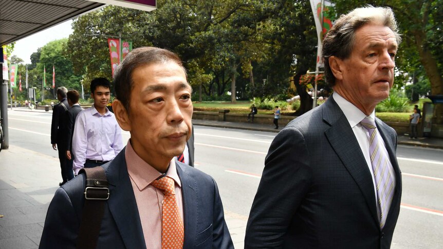 A man wearing a suit and orange tie and shirt walks with another man on a city footpath.