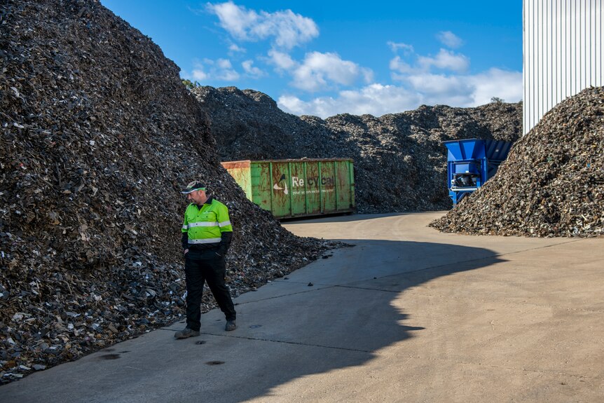A man in a fluro yellow vest walks passed large piles of shredded scrap metal, a green shipment container and blue machinery.