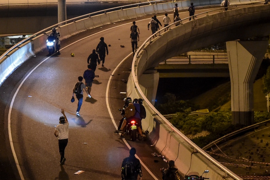 On a highway exit overpass, you see a line of protesters running on it as some motorbikes are waiting to pick them up.