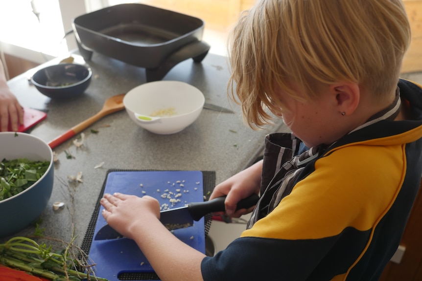 A small boy carefully chops up some garlic on a blue chopping board. He's surrounded by bowls, spoons and an electric fry pan