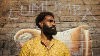 Heritier Lumumba stands in front of a wall with a mural painted on it of Patrice Lumumba.