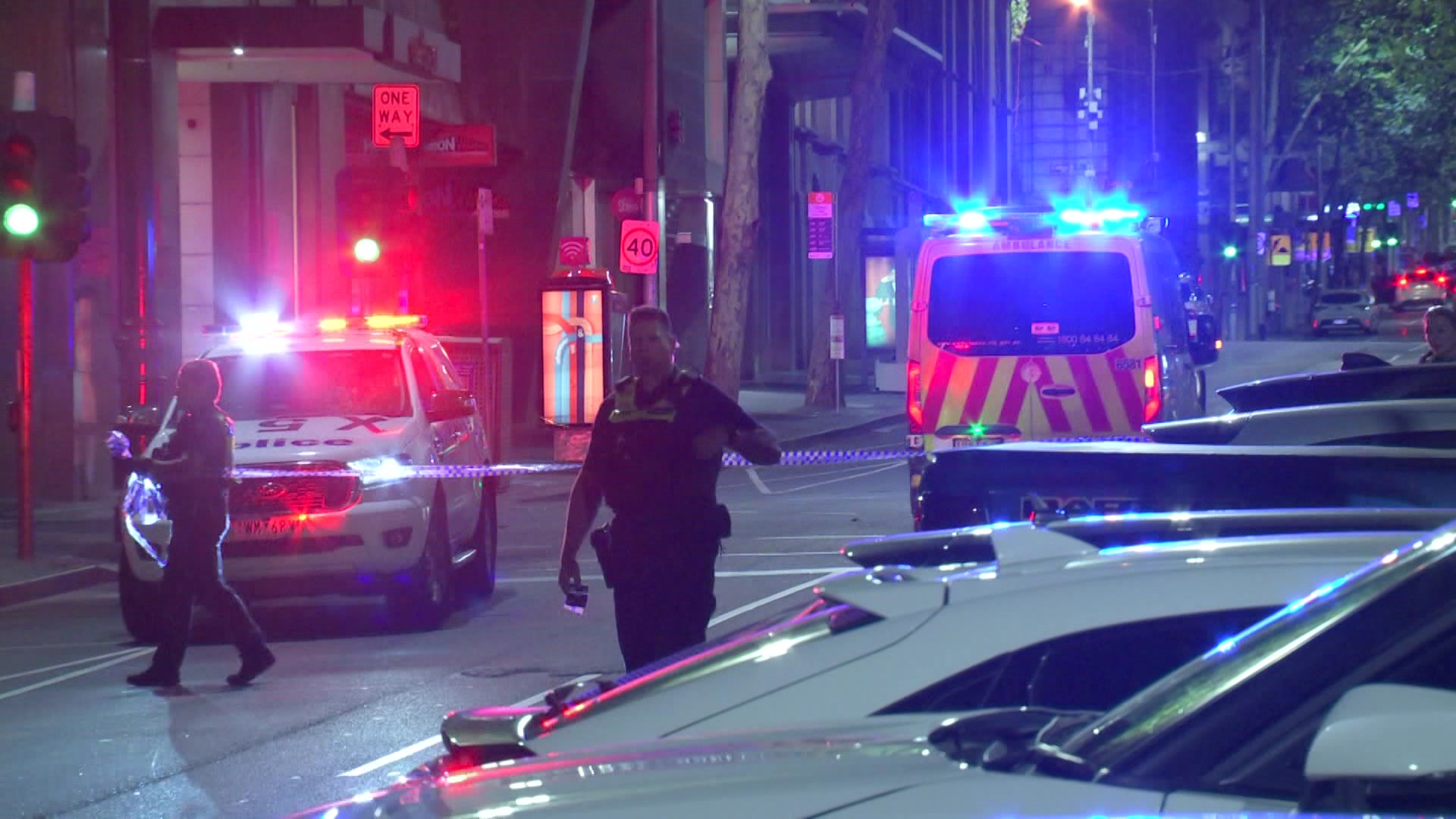 Dos policías caminan por el centro de una calle del centro, detrás de ellos un coche de policía y una ambulancia con las luces encendidas.