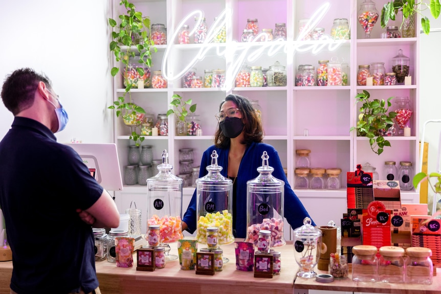 A woman in a mask stands behind a counter covered in candy jars speaking to a customer. 