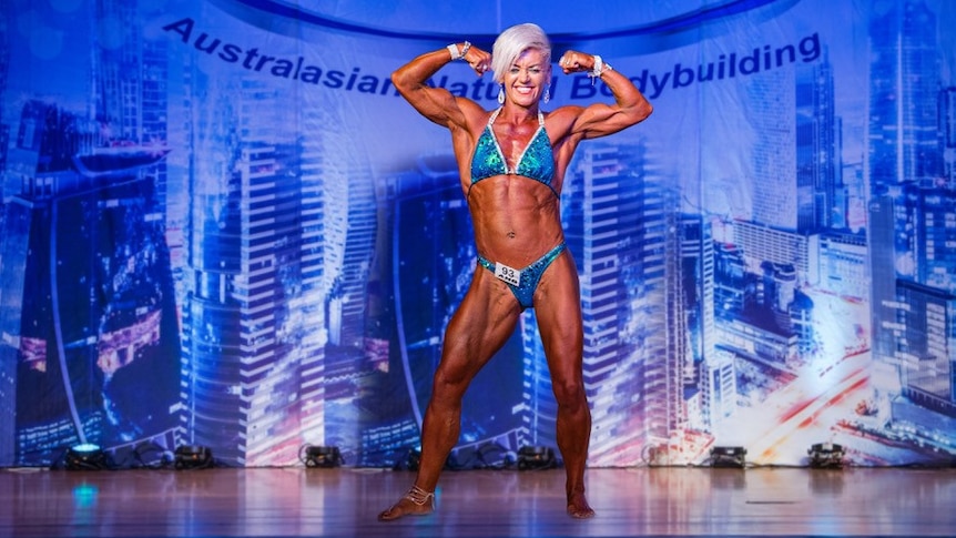 Female bodybuilder stands on stage smiling and arms flexed.