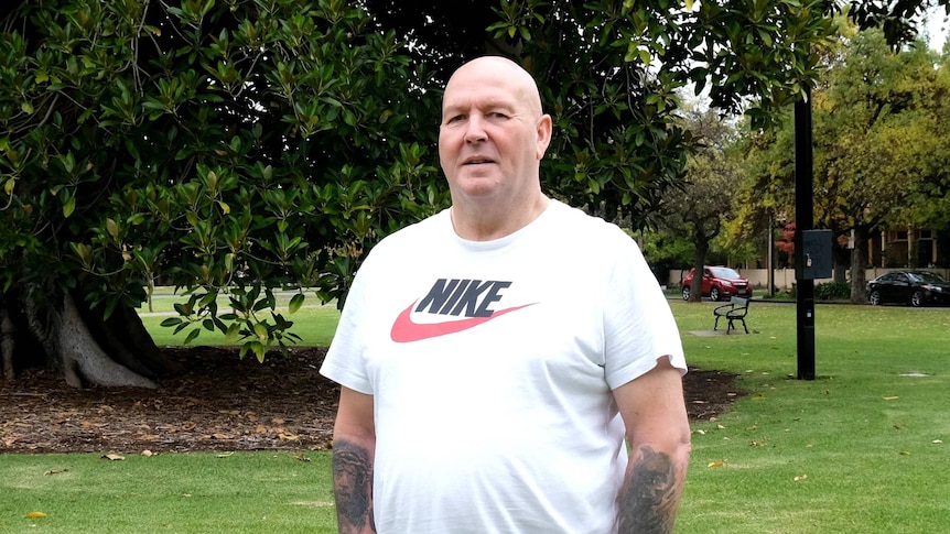 A man in a T-shirt with his hands in a pockets in an urban park.