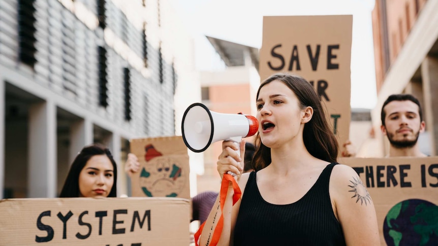 Young woman wearing a black singlet announcing through a megaphone. People behind her hold signs about climate change.