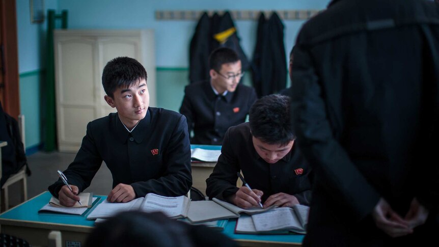 A dim teal classroom shows rows of North Korean school children studying at desks while a teacher paces past them.