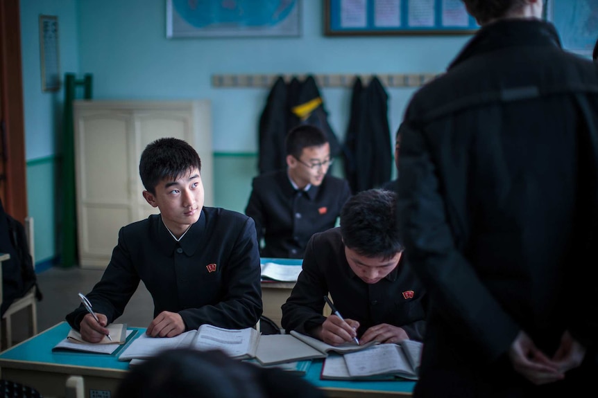A dim teal classroom shows rows of North Korean school children studying at desks while a teacher paces past them.